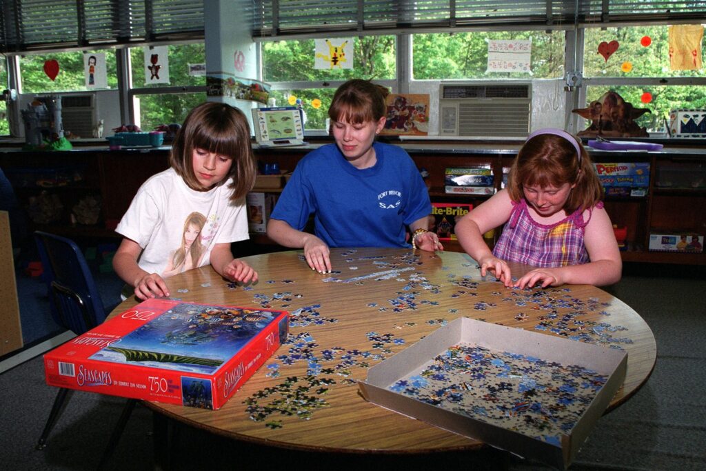Drei Kinder puzzeln gemeinsam an einem runden Tisch mit einer offenen Puzzleschachtel