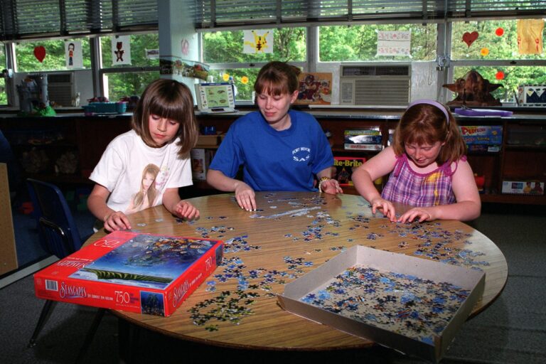 Drei Kinder puzzeln gemeinsam an einem runden Tisch mit einer offenen Puzzleschachtel