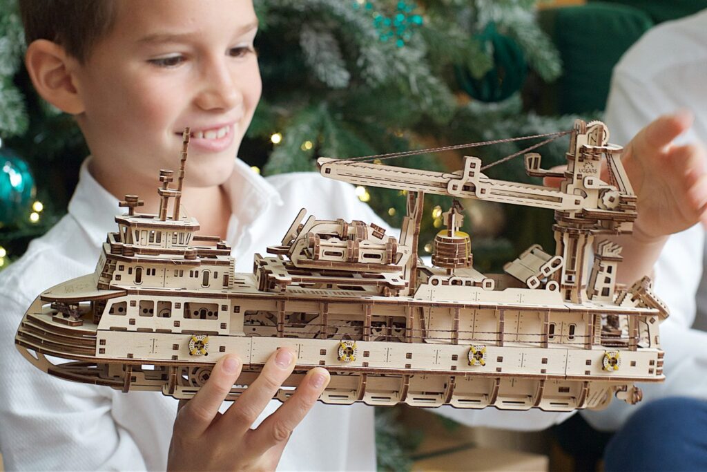 Junge hält ein selbst gebautes Holzmodell eines Schiffes vor einem Weihnachtsbaum